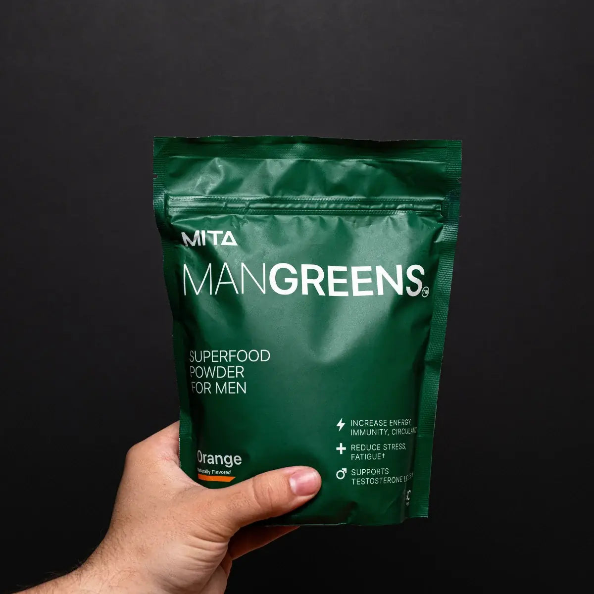 A hand holding a real bag of Man Greens, orange flavor, against a black background